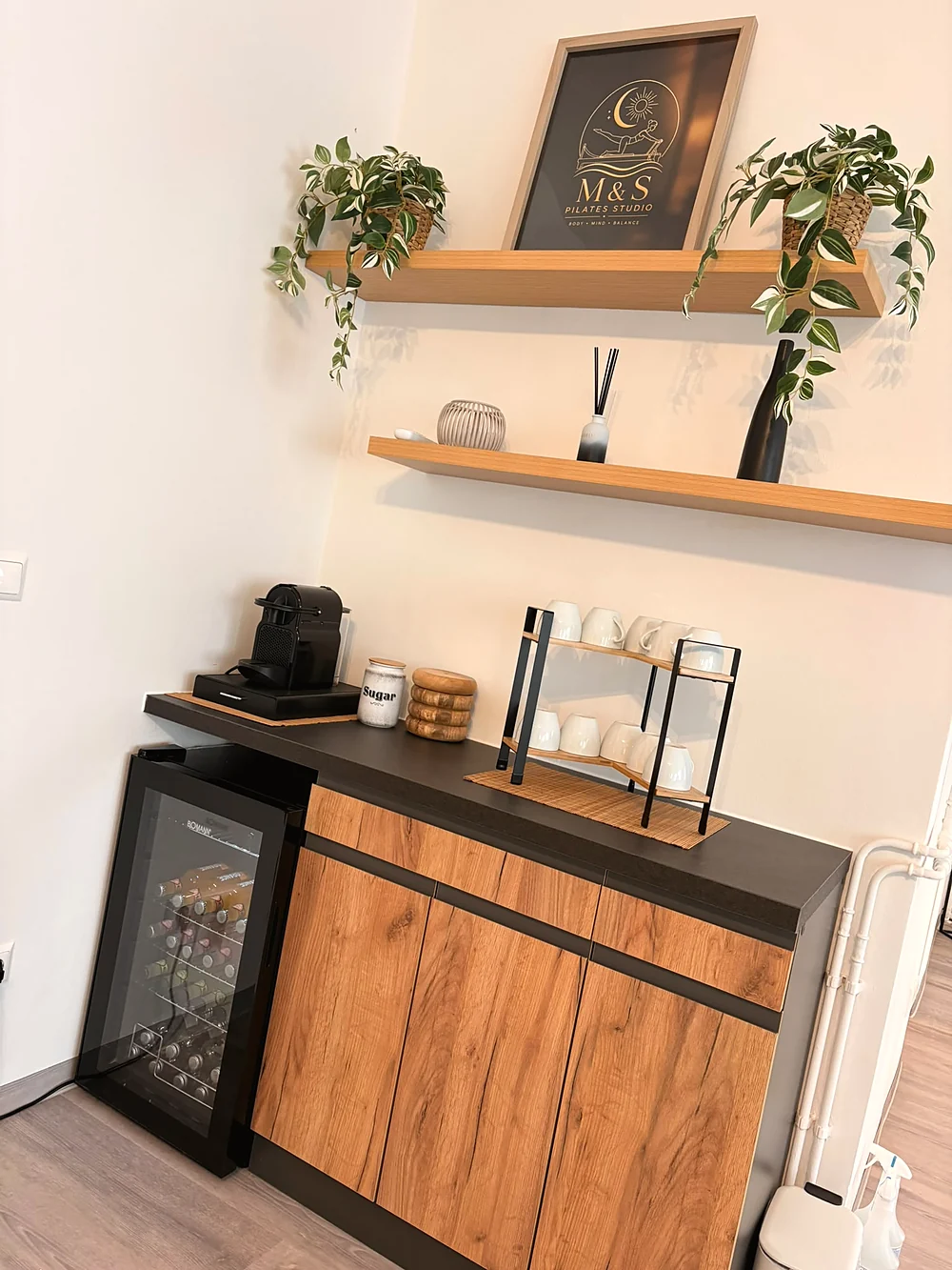 Kaffeeecke mit Kaffeemaschine, Tassen, Getränkekühlschrank und dem gerahmten M&S-Pilates-Studio-Logo im Regal.