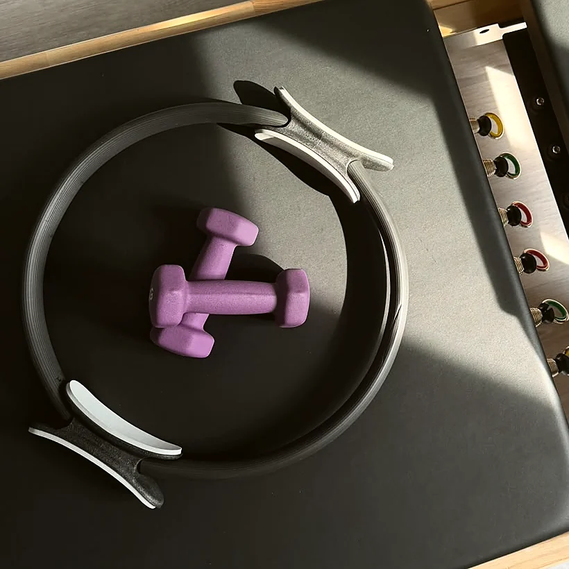 Pilates-Ring und zwei Handgewichte auf dem Reformer, daneben die farbigen Federn.
