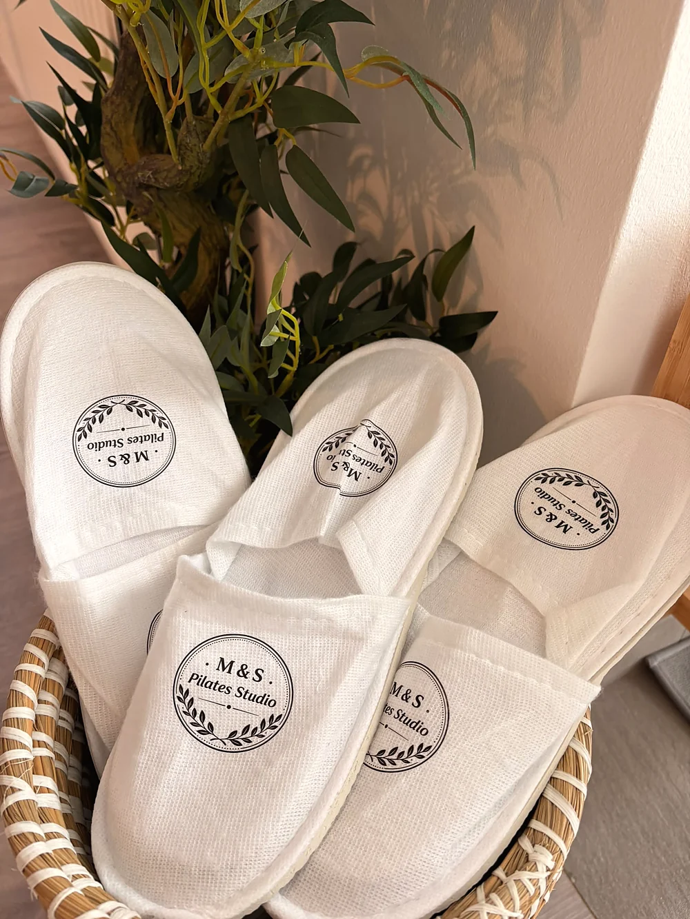 Weiße Slipper mit aufgedrucktem M&S-Pilates-Studio-Logo in einem Korb neben einer Pflanze.